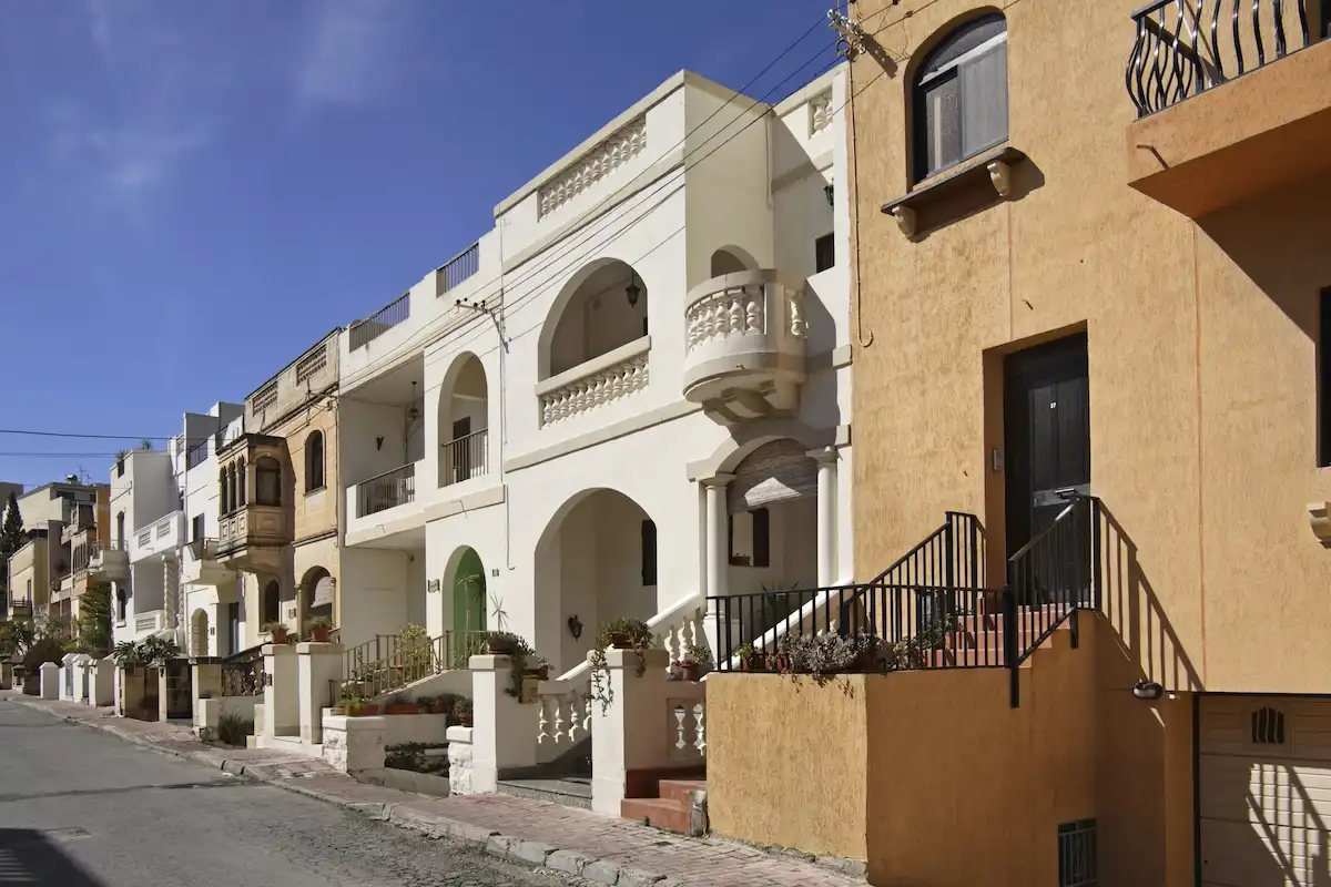 Row of typical Maltese terraced houses on a quiet residential street with balconies and staircases under a clear blue sky.