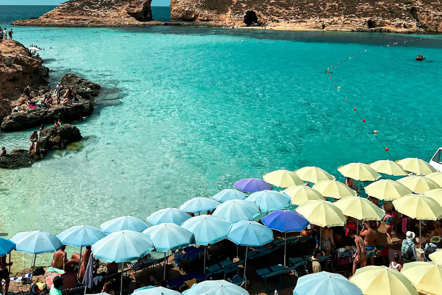 Overcrowded Blue Lagoon on Comino, Malta, showing rows of sunbeds and umbrellas beside turquoise water in peak summer