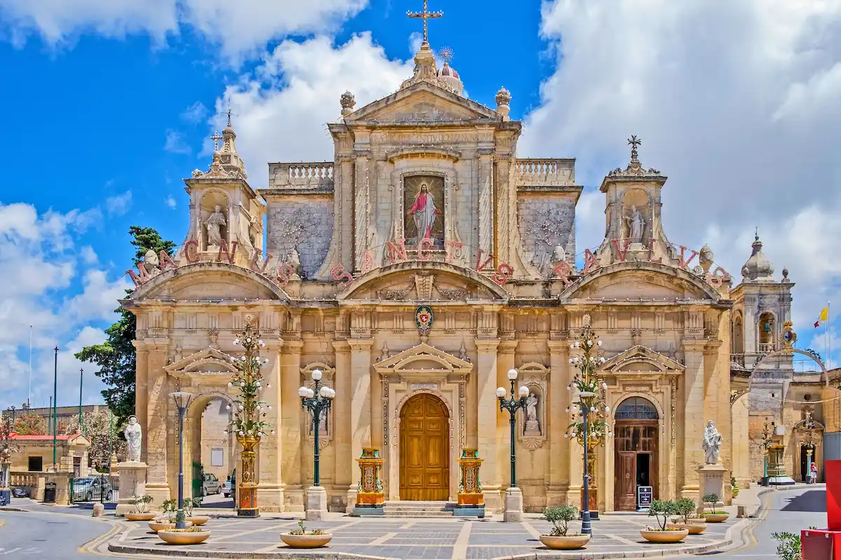 Baroque Collegiate Church of St Paul in Rabat, Malta, with an ornate limestone façade, statues of saints, a central wooden portal, and a cross at the top, photographed on a sunny day with a blue sky and scattered clouds.