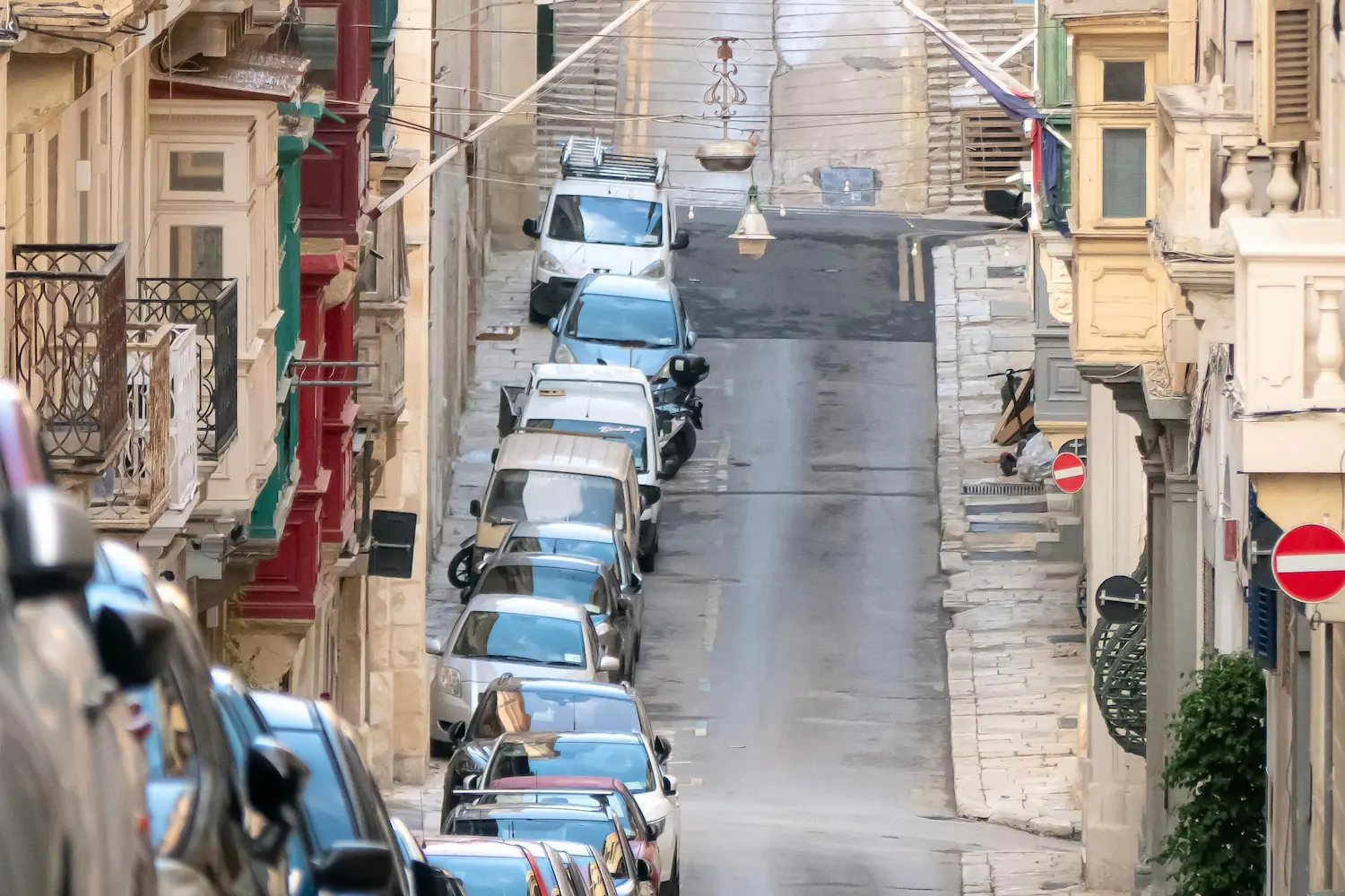 Narrow residential street in Malta with parked cars and traditional balconies, illustrating everyday housing and transport conditions.